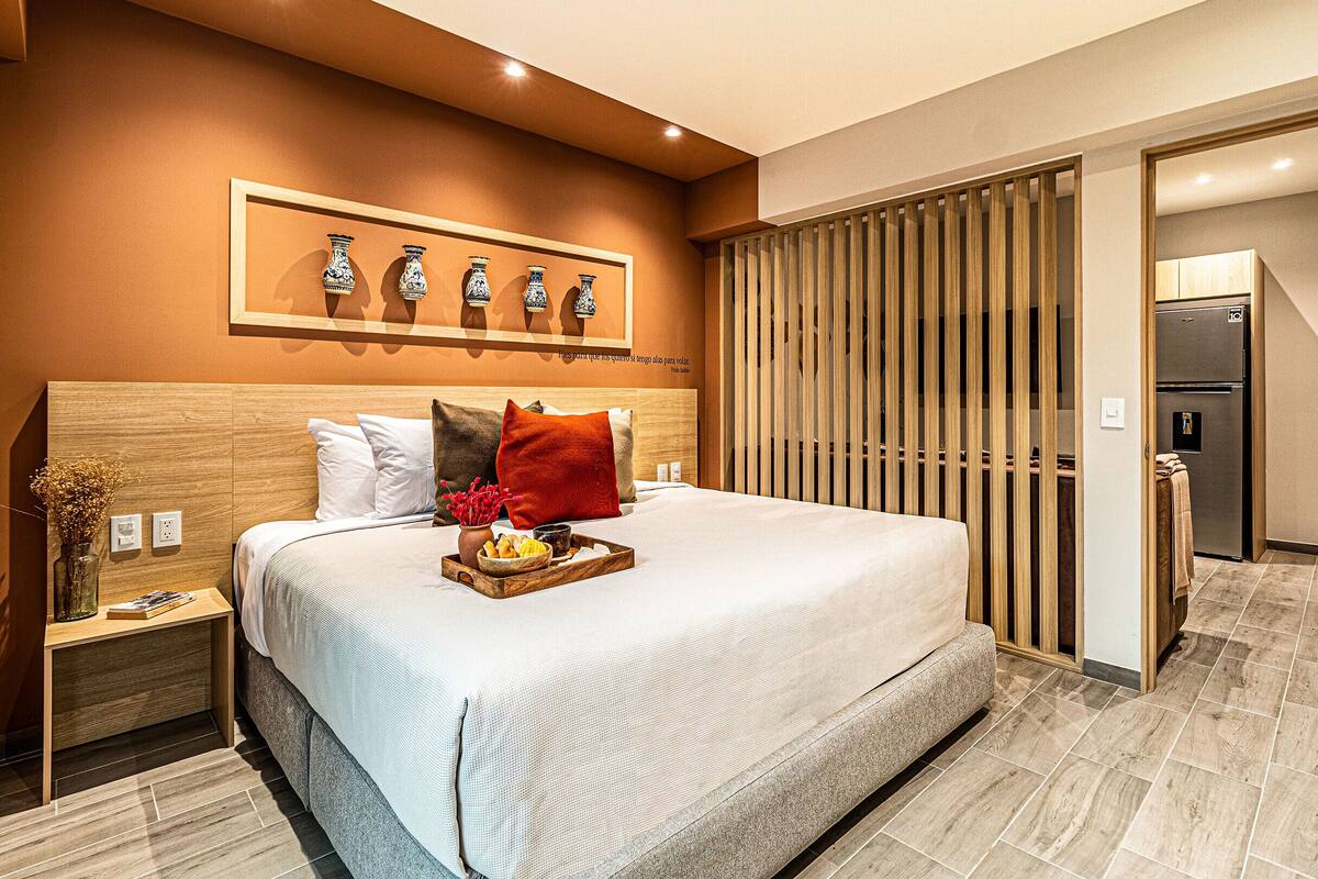 Habitació d'hotel moderna amb un llit gran, un capçal de fusta, una tauleta de nit amb un vas i un llibre, i una bandeja de petit esmorzar al llit.