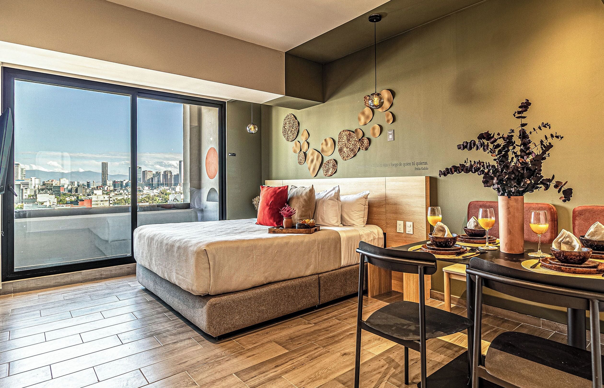 Habitación de hotel moderna con una cama grande, una mesa de comedor con sillas y vista a la ciudad a través de un ventanal amplio.