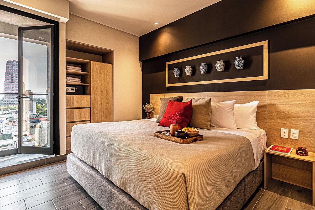 A modern hotel room with a large bed, a wooden headboard, a nightstand, and a view of the city through a large window.