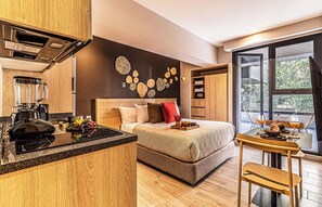 Une chambre d’hôtel moderne dotée d’un coin cuisine, d’un lit avec une tête de lit, d’une table à manger avec des chaises et d’une grande fenêtre offrant une vue dégagée.