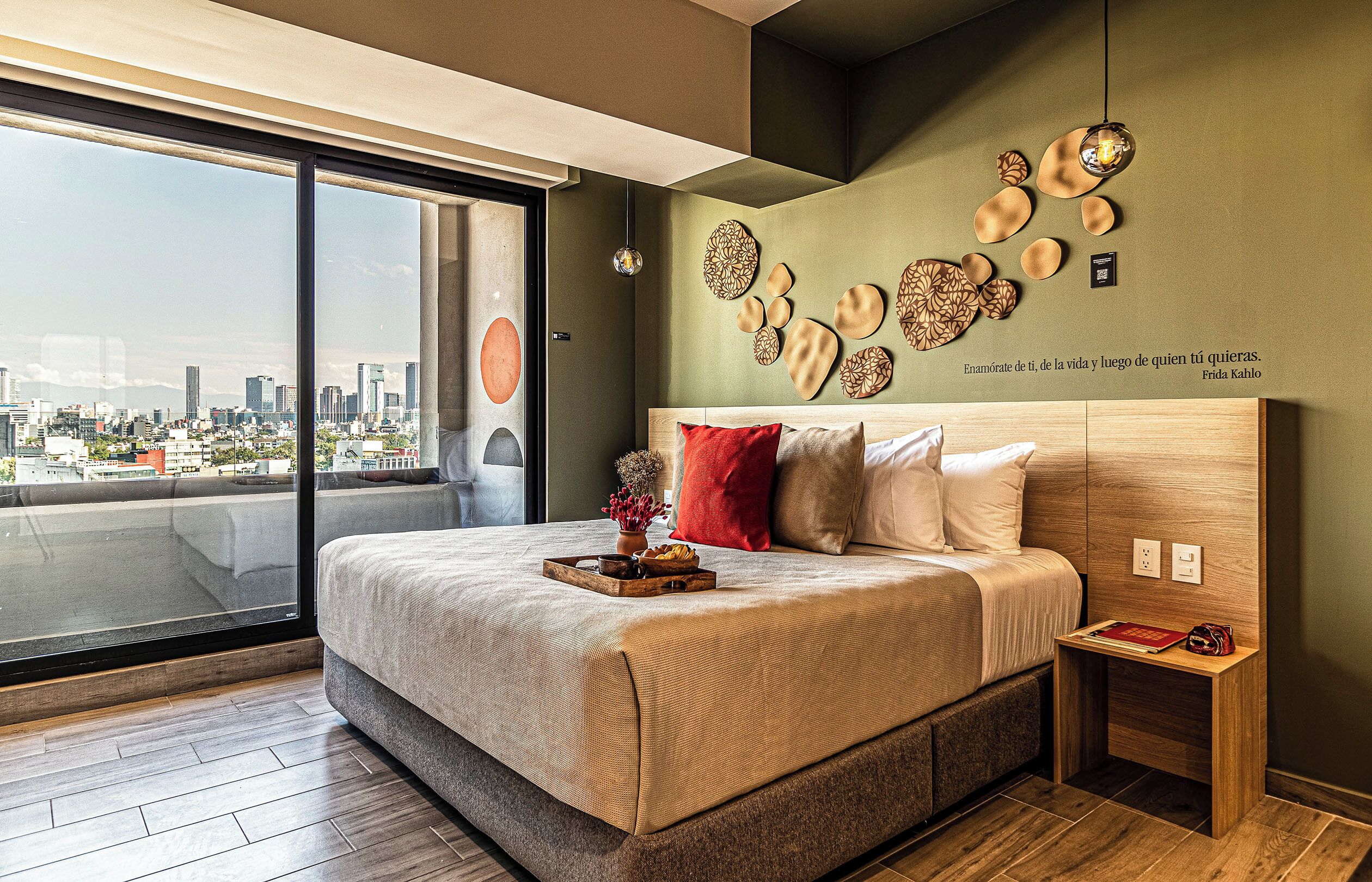 Quarto moderno com vista para a cidade, uma cama grande, arte decorativa na parede e uma mesa de cabeceira com um livro e uma bolsa.