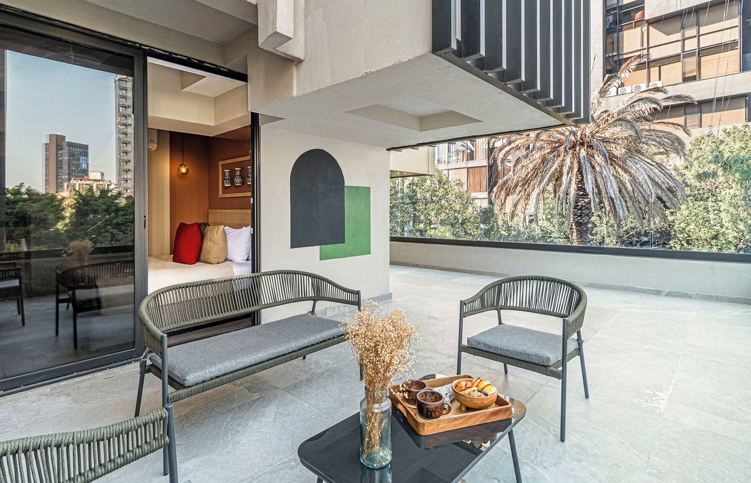 Un área de estar moderna al aire libre con muebles de mimbre, una mesa de vidrio con una bandeja de bocadillos y vistas a una palmera y edificios.