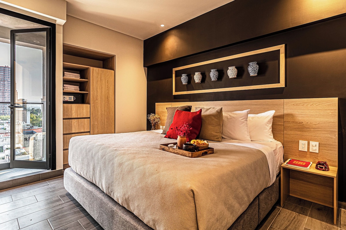 Una habitación de hotel moderna con una cama grande, cabecera de madera, mesita de noche y vista a la ciudad a través de un ventanal.