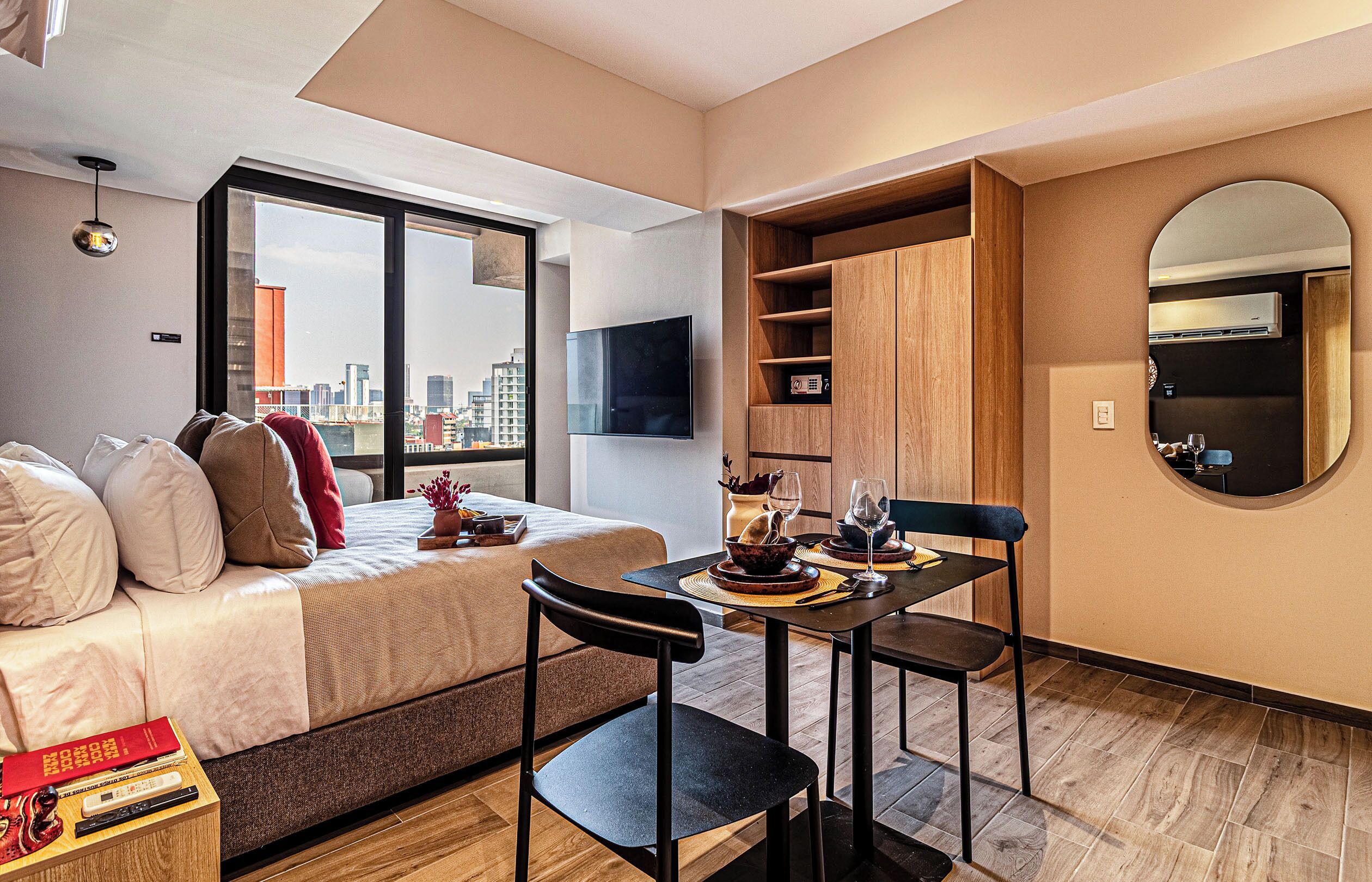 A modern hotel room with a bed, a dining table with chairs, a small kitchen area, and a large window offering a city view.