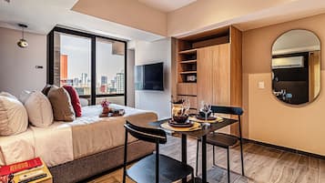 Une chambre d’hôtel moderne comprenant un lit, une table à manger avec des chaises, un petit coin cuisine et une grande fenêtre donnant sur la ville.