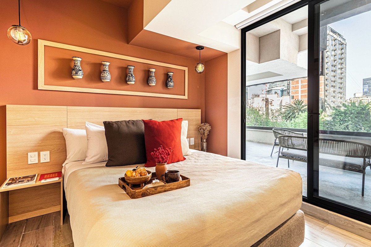Habitación de hotel con una cama grande, cabecera de madera, vistas de la ciudad a través de un ventanal y un balcón con mobiliario exterior.