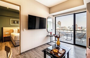 Une chambre d’hôtel comprenant un lit, une télévision, une table à manger avec un vase et un panier de pain, ainsi qu’un balcon avec une chaise et une table.