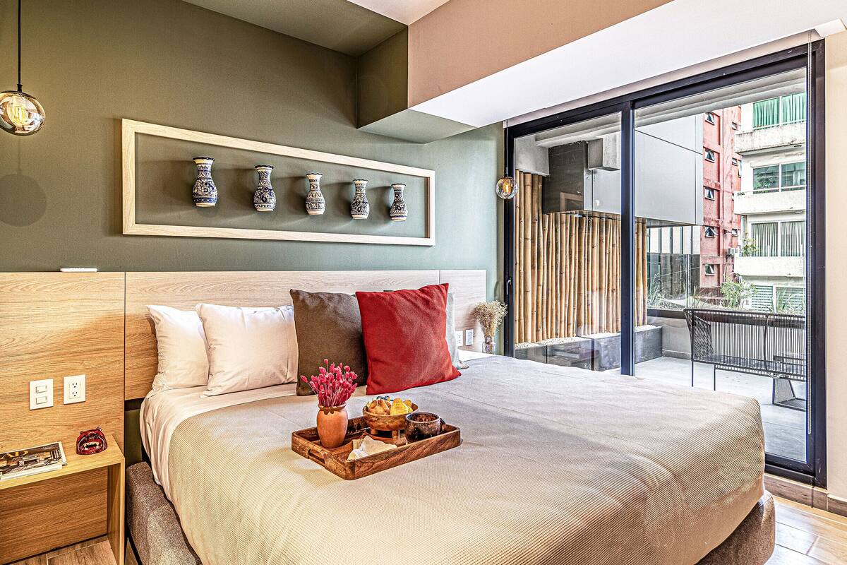 Habitació d'hotel moderna amb un llit gran, una petita taula de nit de fusta, un safata amb aperitius i un balcó amb vistes a edificis.