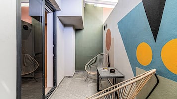 Un pasillo con una puerta de cristal que da a una habitación, una pequeña mesa con una revista, dos sillas de mimbre y una pared con motivos geométricos abstractos.