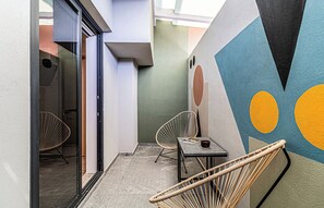 Un pasillo con una puerta de cristal que da a una habitación, una pequeña mesa con una revista, dos sillas de mimbre y una pared con motivos geométricos abstractos.