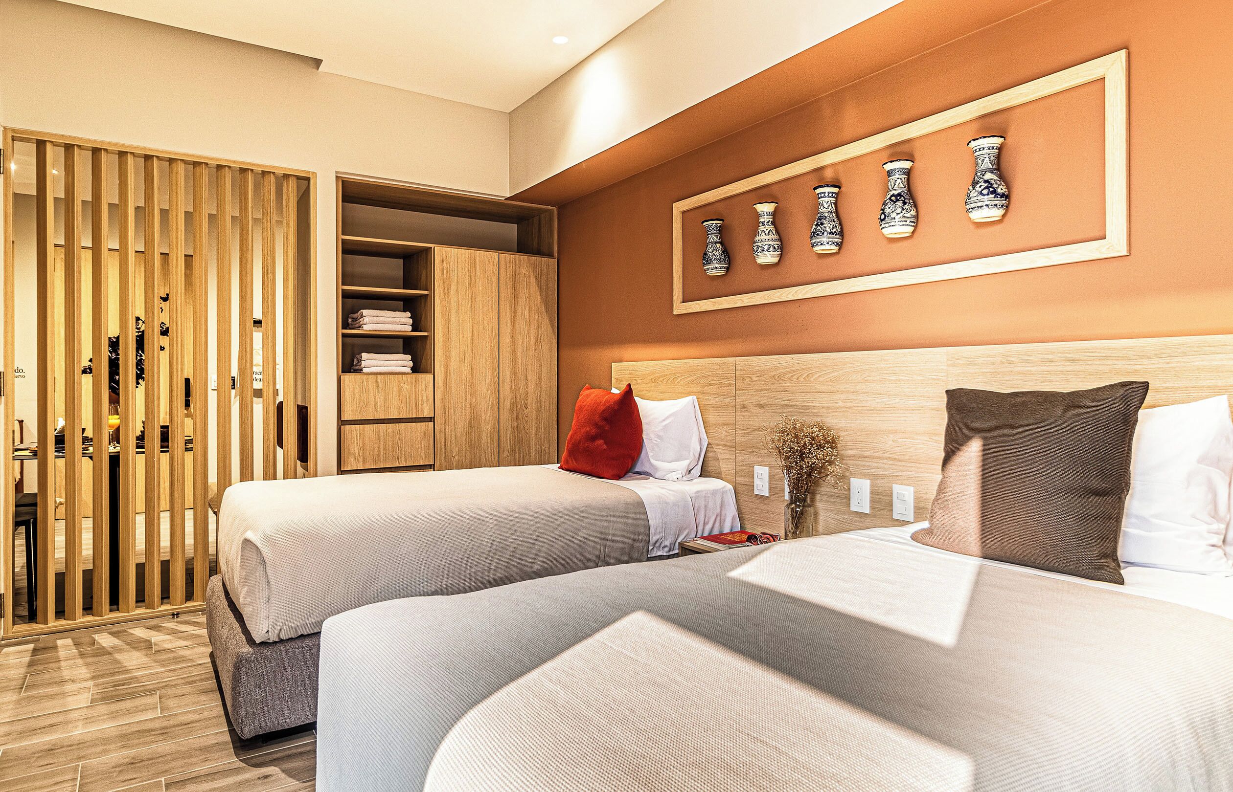 A hotel room with two beds, a wooden wardrobe, a wall-mounted vase display, and a small table with a vase and book.