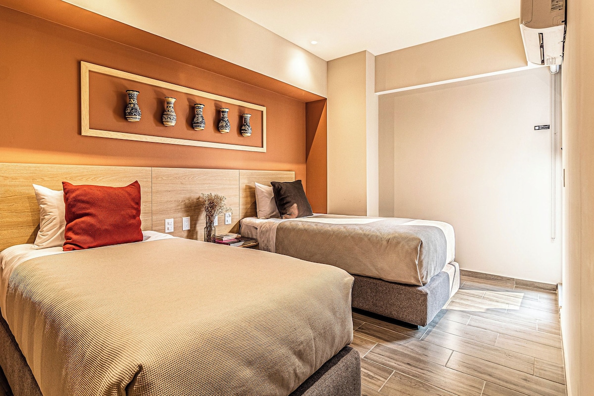 Habitación de hotel con dos camas, cabecero de madera, un jarrón decorativo en la pared y una puerta con pomo.