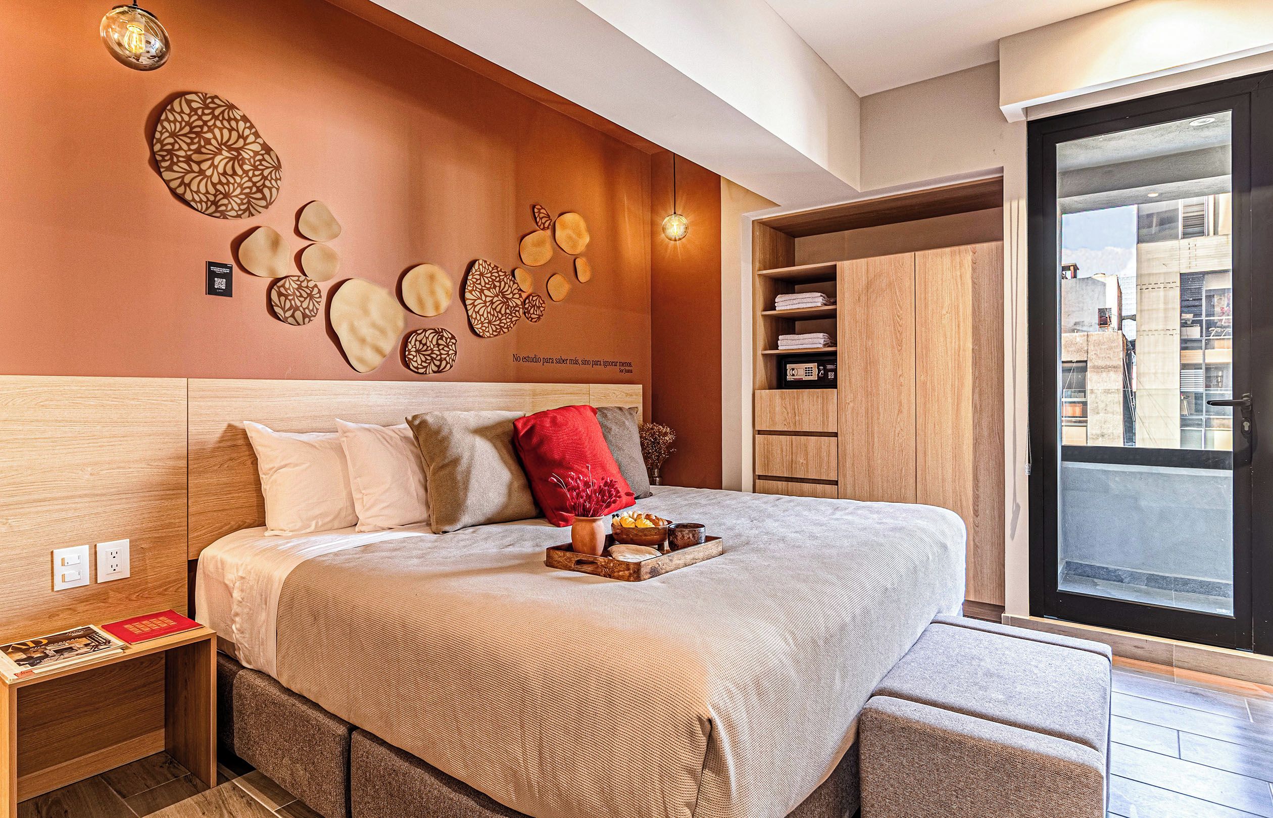 Una habitación de hotel moderna con una cama grande, una mesita de noche, un cojín decorativo rojo y vistas a la ciudad a través de un ventanal amplio.