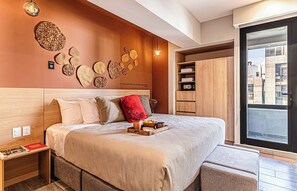 Una habitación de hotel moderna con una cama grande, una mesita de noche, un cojín decorativo rojo y vistas a la ciudad a través de un ventanal amplio.