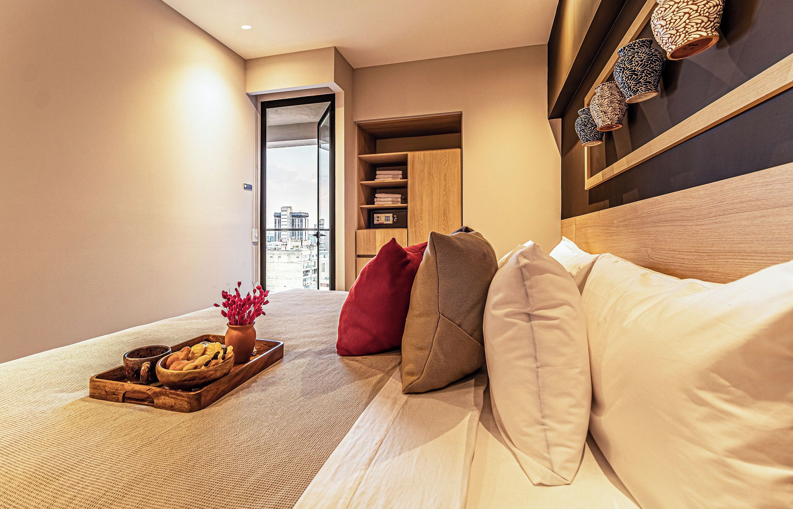 Una habitación de hotel moderna con cama, mesita de madera con bandeja de comida y flores, armario y vistas a la ciudad a través de una ventana.