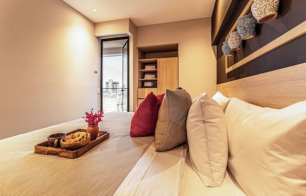 Une chambre d’hôtel moderne avec un lit, une table de chevet en bois sur laquelle se trouve un plateau avec de la nourriture et des fleurs, une armoire et une vue sur la ville par la fenêtre.