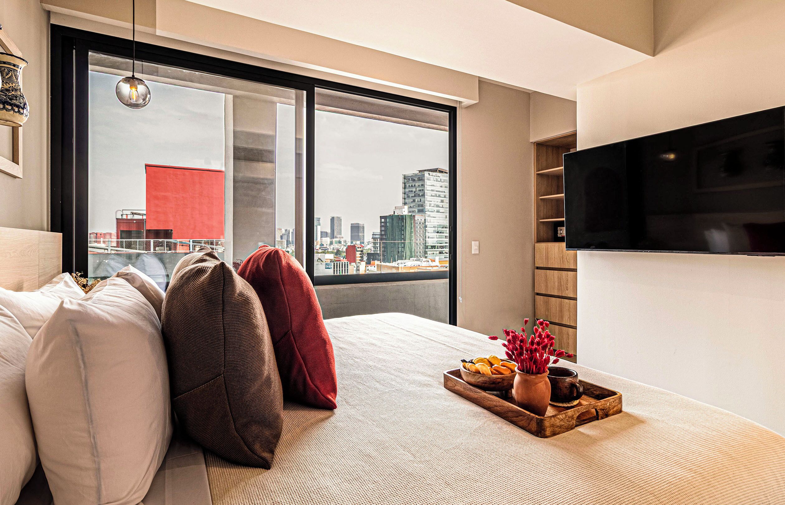 A modern bedroom with a large window, a bed with pillows, a tray with bread and flowers, and a TV on the wall.