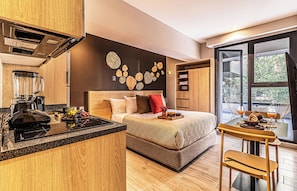 Une chambre d’hôtel moderne dotée d’un coin cuisine, d’un lit avec une tête de lit, d’une table à manger avec des chaises et d’une grande fenêtre offrant une vue dégagée.