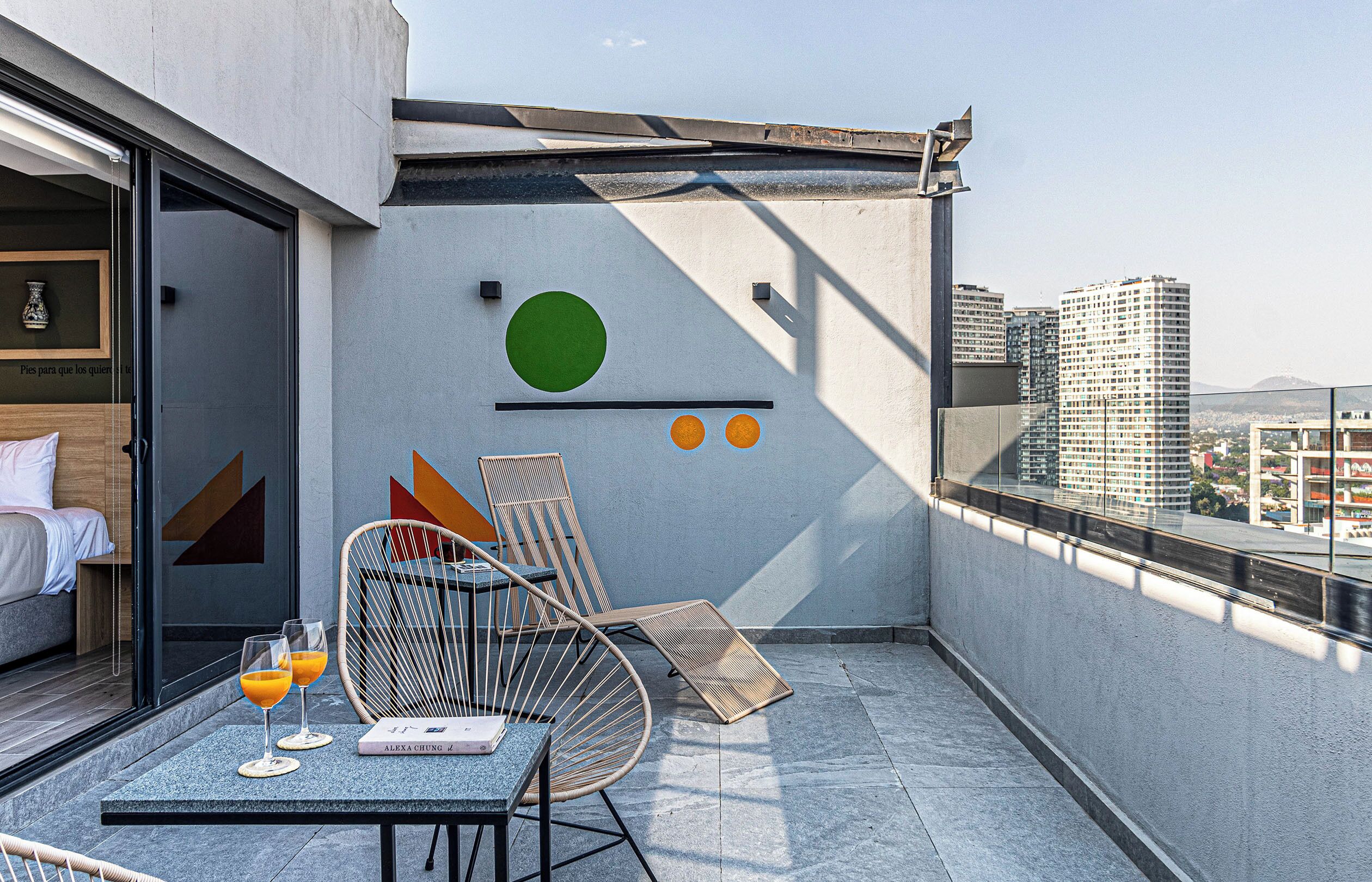 Un balcón moderno con vista a la ciudad, una silla de mimbre, una mesita con bebidas y un libro.