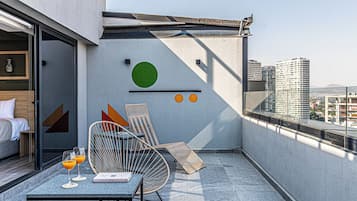 Un balcón moderno con vista a la ciudad, una silla de mimbre, una mesita con bebidas y un libro.