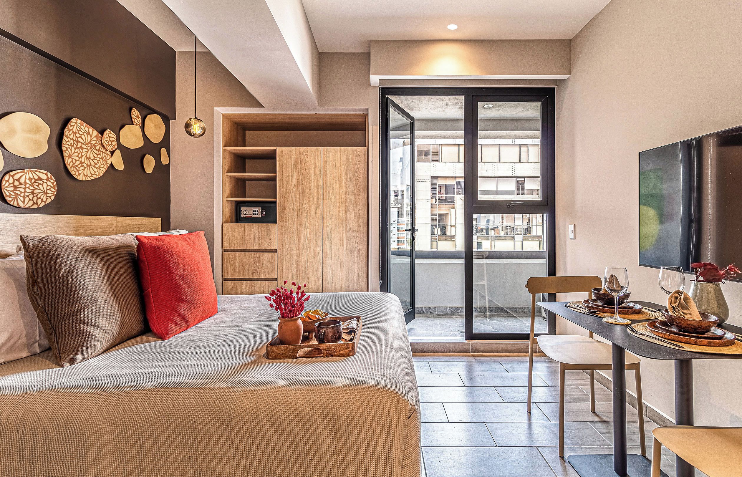 Une chambre moderne avec un lit, un coin repas avec une table dressée pour deux personnes, et une porte-fenêtre coulissante donnant sur un balcon.