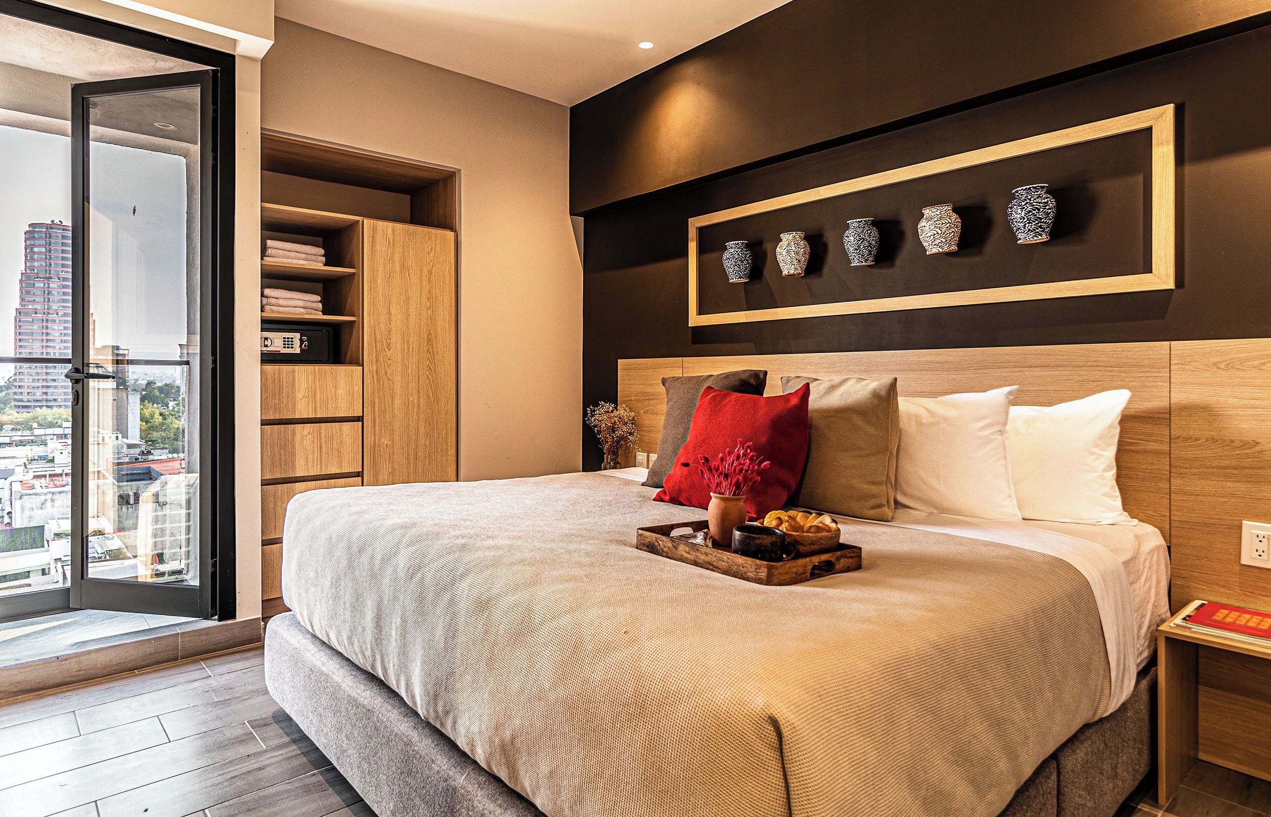 A modern bedroom with a large bed, a wooden wardrobe, a nightstand, and a city view through a glass door.