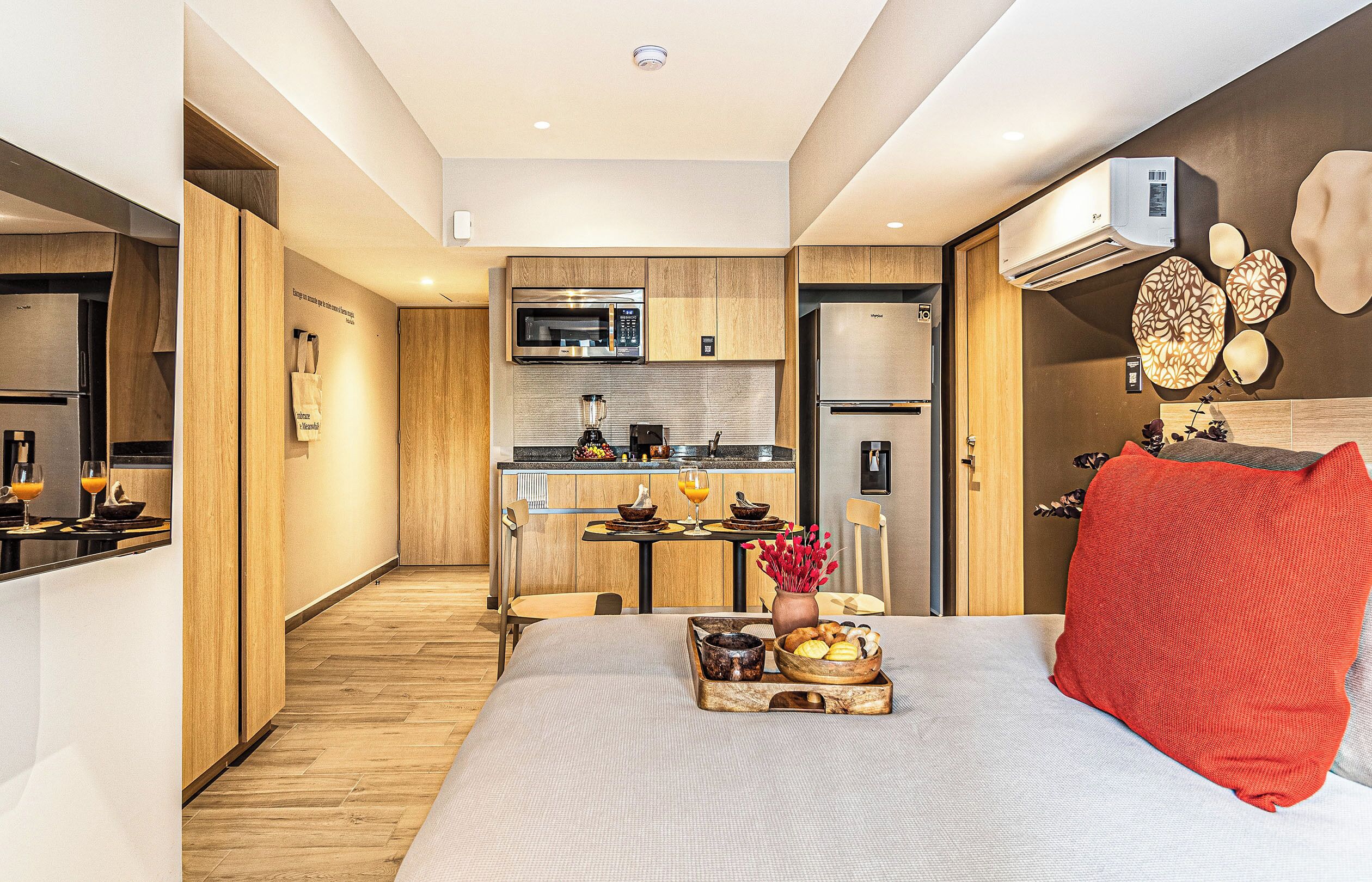 Una habitación de hotel moderna con kitchenette, una cama con almohada roja y una bandeja de desayuno sobre la cama.