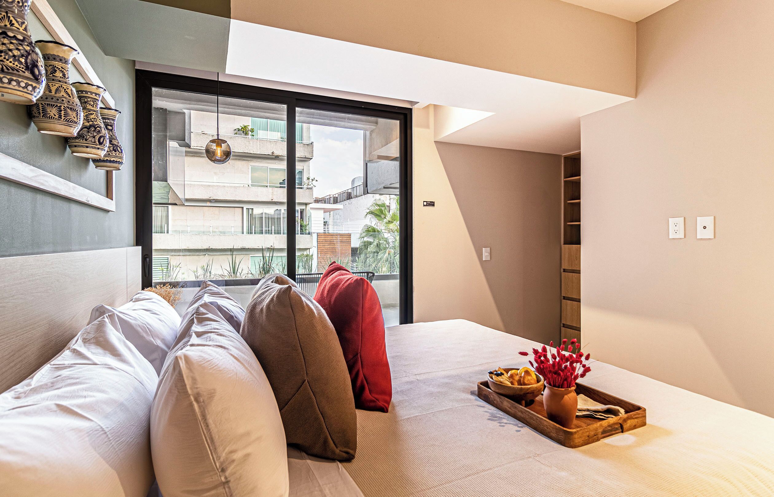 Habitación de hotel moderna con una cama, almohadas, una bandeja con comida y flores, una ventana con vistas a edificios y un estante de pared con adornos.