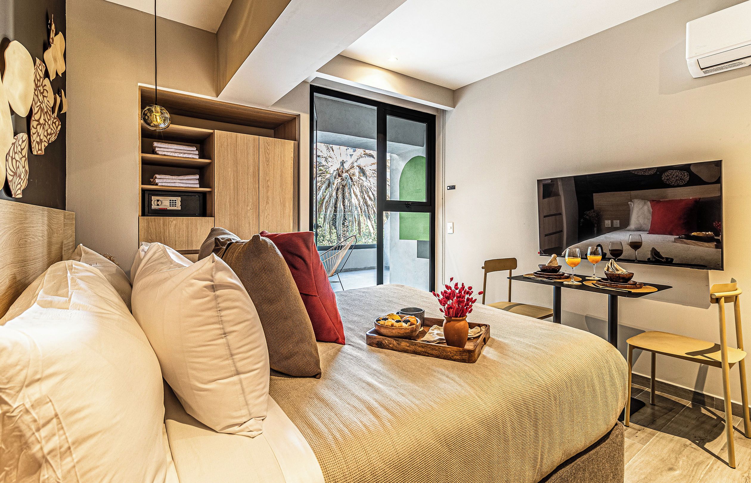 A modern bedroom with a large bed, a TV, a dining area, and a view of palm trees.