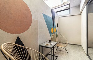 Un interior moderno con un mural geométrico en la pared, dos sillas de mimbre, una mesita con una revista y un tragaluz.