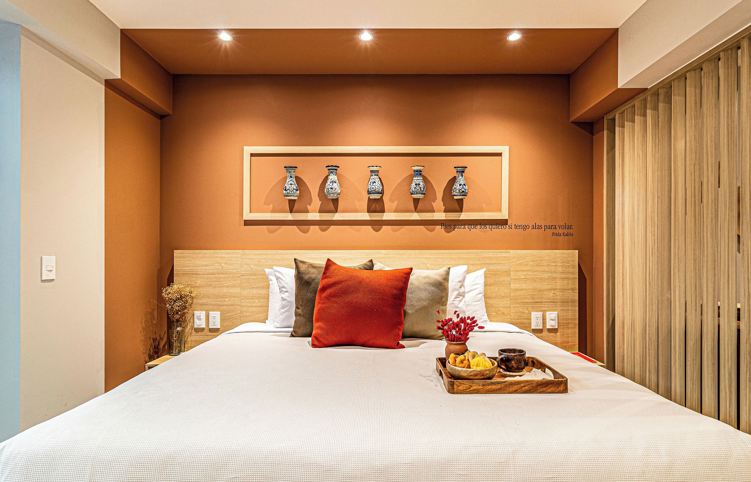 A hotel room with a large bed, a wooden headboard, a wall-mounted shelf with decorative vases, and a tray with bread and a cup on the bed.