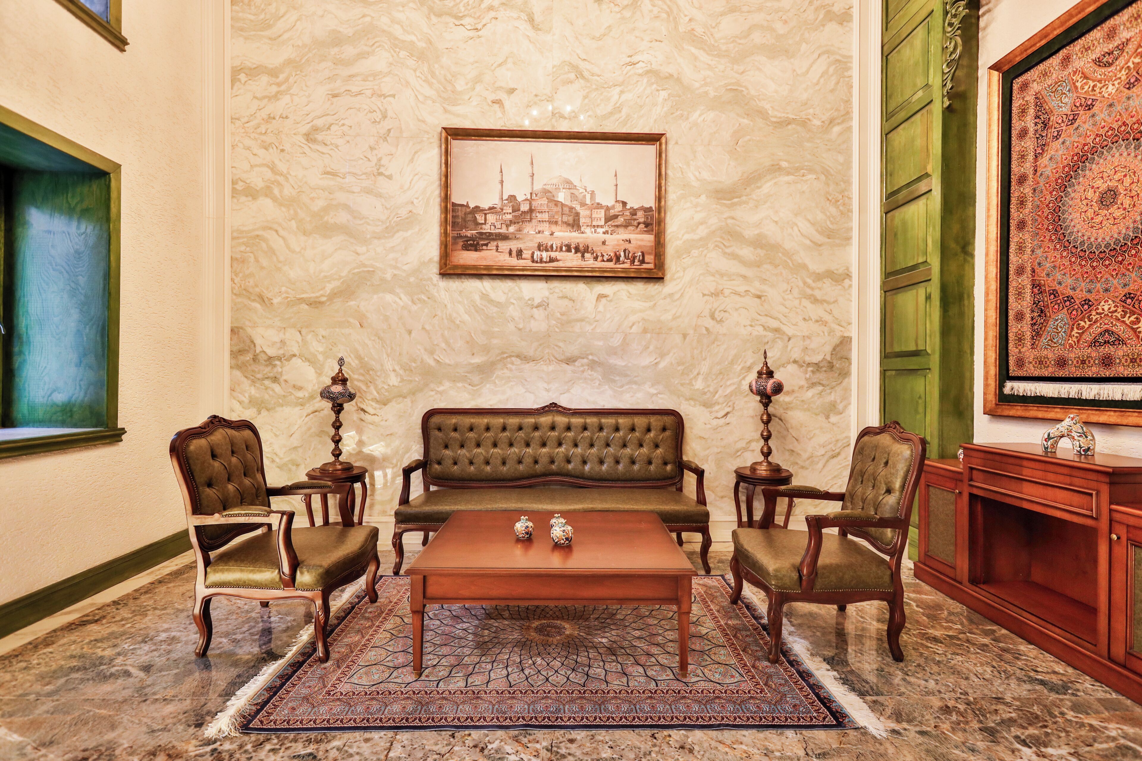 Una habitación con un sofá verde, dos sillones, una mesa de centro de madera, una alfombra estampada, un cuadro en la pared y una puerta verde.