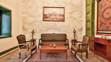 Una habitación con un sofá verde, dos sillones, una mesa de centro de madera, una alfombra estampada, un cuadro en la pared y una puerta verde.