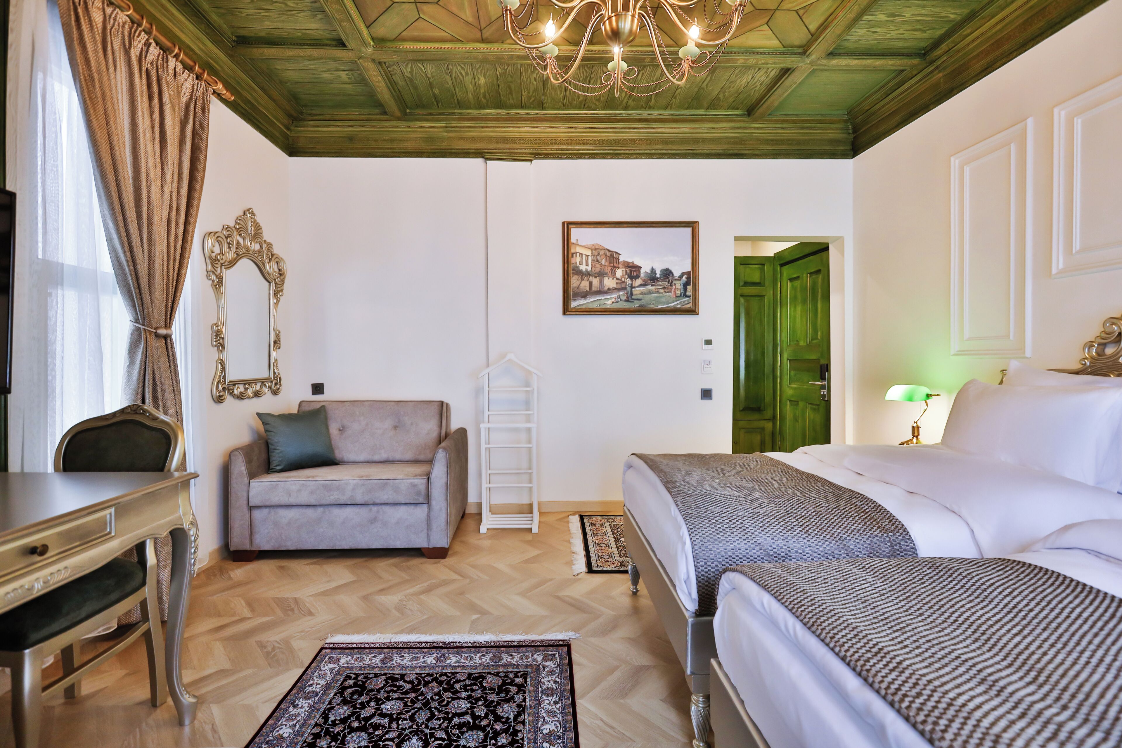 A hotel room with a bed, a sofa, a desk, a chandelier, and a painting on the wall.