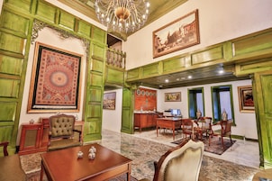 A spacious room with a large ornate chandelier, green paneled walls, and a traditional wooden desk with chairs.
