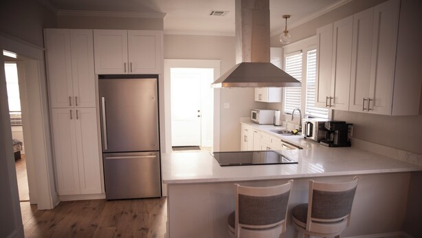 A modern kitchen with white cabinetry, a stainless steel refrigerator, a microwave, and a large island with bar stools. - Charming Meets Modern 2-bedroom Cottage (Morgan City)