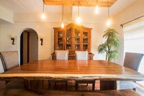 Uma área de refeições com uma grande mesa de madeira, cadeiras, um armário com portas de vidro, luminárias pendentes e uma planta.