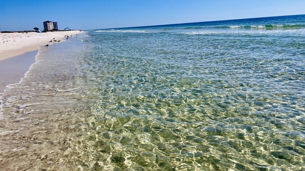 Una playa con aguas transparentes y poco profundas, una orilla arenosa y un edificio a lo lejos.