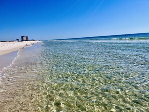 Ein Strand mit klarem, seichten Wasser, einem sandigen Ufer und einem Gebäude in der Ferne.