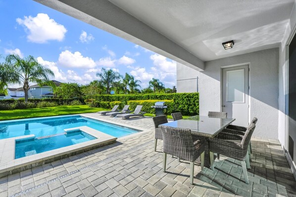 A pool area with a glass table and wicker chairs, a barbecue grill, and a view of a green lawn and trees.