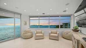 A spacious room with large windows offering a view of the sea and a city skyline. There are two beige armchairs and a wooden sideboard with a plant. - Endless Horizon in Siesta Key (Siesta Key)