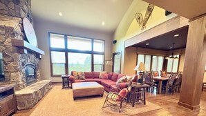 A spacious living room with a stone fireplace, large windows, a sectional sofa, and a dining area with wooden chairs and a table. - 3 Bedroom w/ Game Room / Gourmet Kitchen / Community Pool - MNT VIEW IS AMAZING! (Cle Elum)
