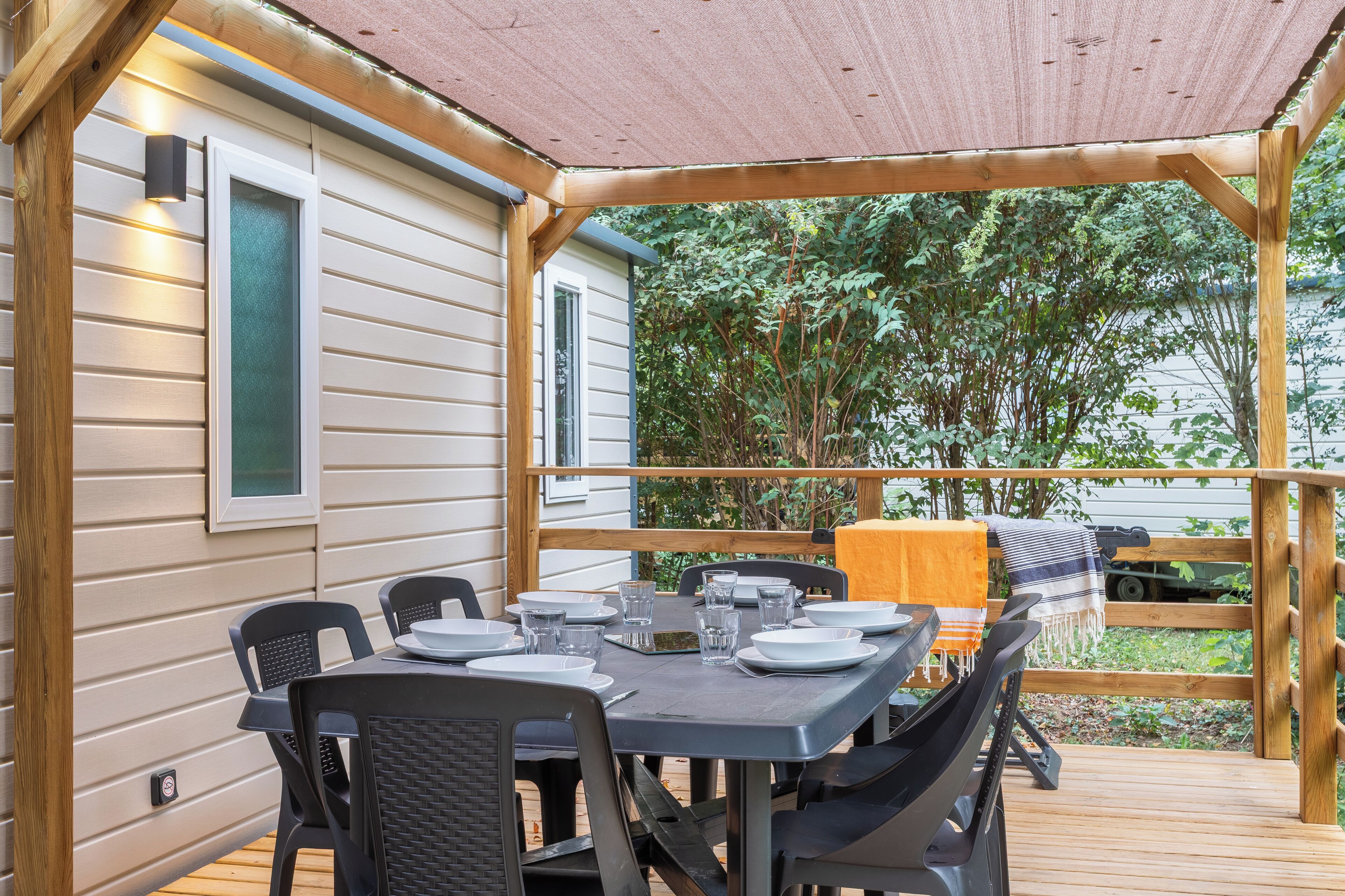Un espacio de comedor al aire libre techado, con una mesa dispuesta para cuatro personas, sillas alrededor y una terraza de madera con vistas a la vegetación.