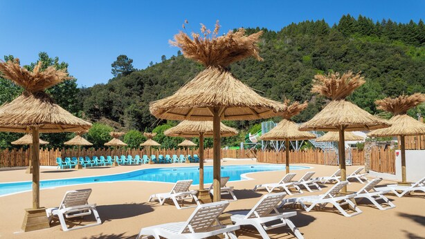 Un espace piscine doté de chaises longues et de parasols en chaume, entouré d’une clôture en bois et bordé d’une colline boisée.