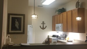 Una cocina con armarios de madera, microondas y encimera con diversos objetos. Hay dos lámparas colgantes, una luz de techo e imagen enmarcada en la pared.