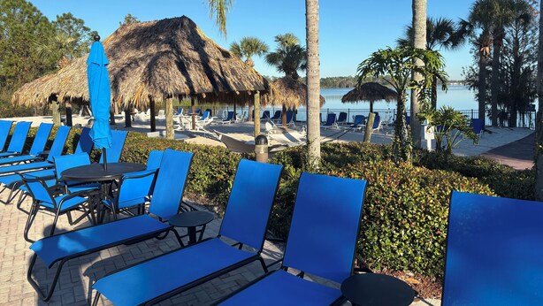 Un área de descanso junto a la playa con sillas y mesas azules, una estructura de techo de paja y vista al agua.