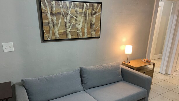 A light gray sofa in a room with a textured wall, a framed abstract painting, a small wooden side table with a lamp, and a tiled floor. - Lenny's Southy Hostel (St. Petersburg)