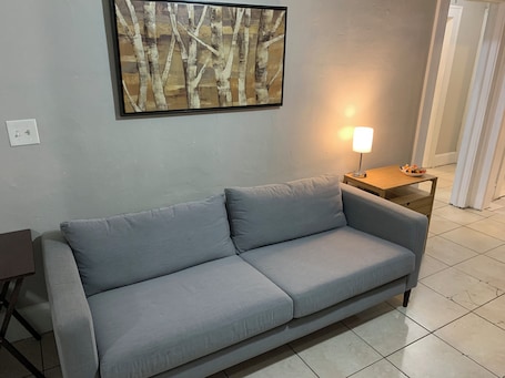 A light gray sofa in a room with a textured wall, a framed abstract painting, a small wooden side table with a lamp, and a tiled floor.