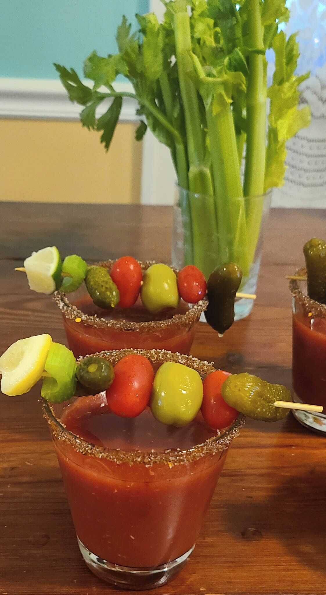 Zwei Bloody Mary Cocktails, garniert mit Oliven und Gewürzgurken, dazu Selleriestangen und Limetten.