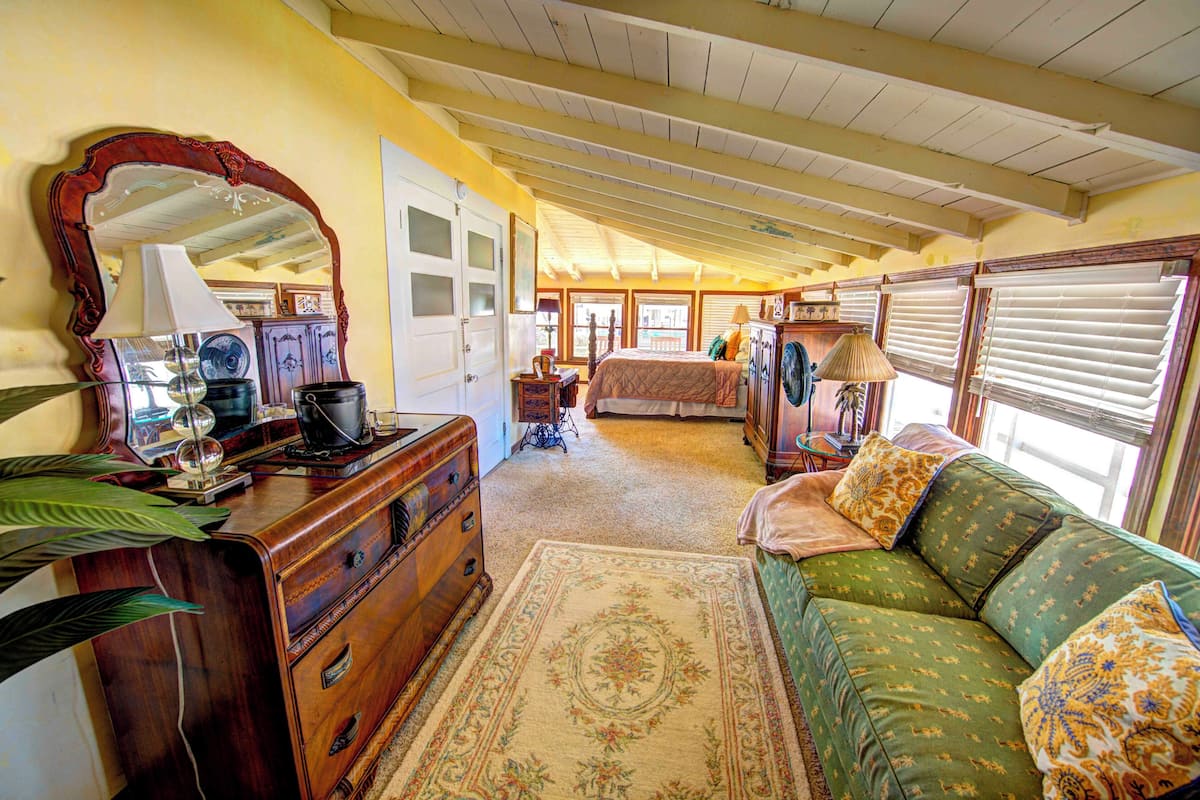 A room with a wooden dresser, a green sofa, a large mirror, and a bed with a floral bedspread.